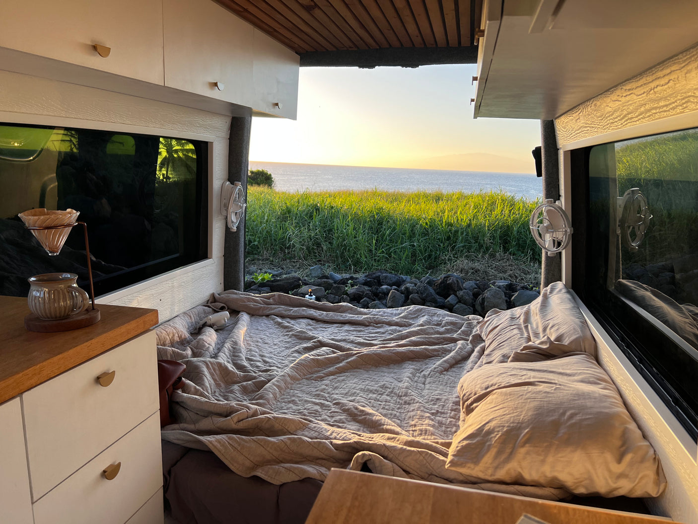 RV rental comfort: A cozy van interior with a bed, coffee setup, and stunning ocean sunset views from the window.