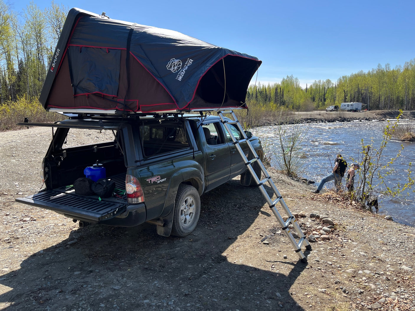 Truck with rooftop tent camped by a river; people and a dog enjoy the bank. An RV rental is visible across the water.