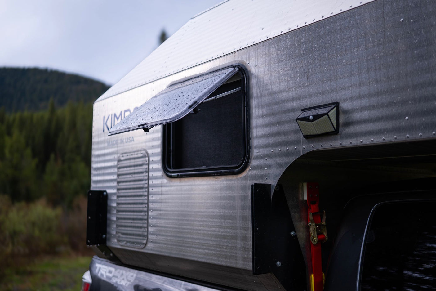 A silver Kimbo truck camper, perfect for RV rental trips, shows its rugged design, open window with raindrops, and solar light.