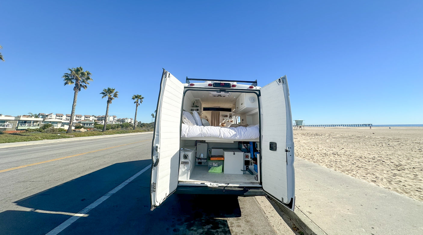 Rv rental parked beachside, rear doors open revealing an elevated bed and storage. Ideal for ocean views on a sunny day.