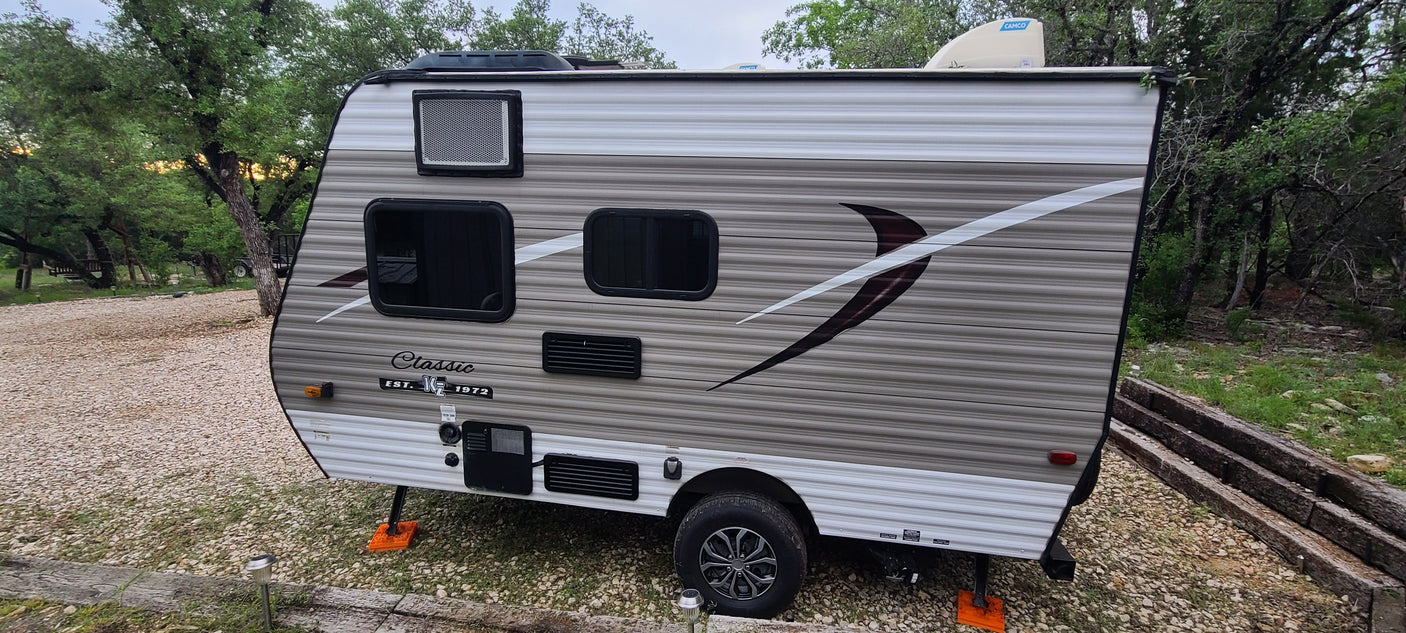 Cozy travel trailer RV rental parked on a gravel campsite amidst lush trees, ready for your next outdoor adventure.