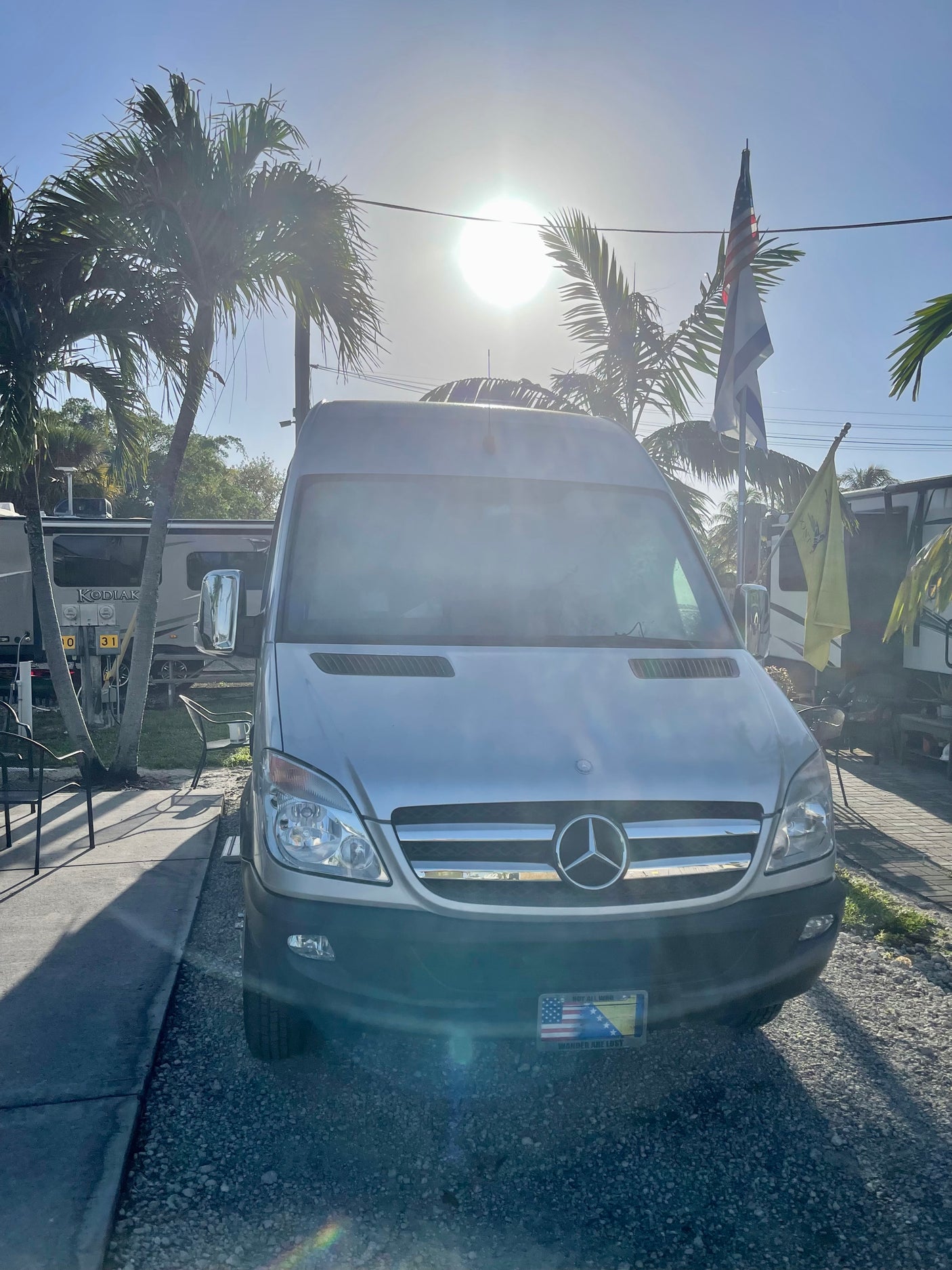A silver Mercedes Sprinter van, a versatile RV rental, parked under sunny skies at a campground with palm trees and other RVs.