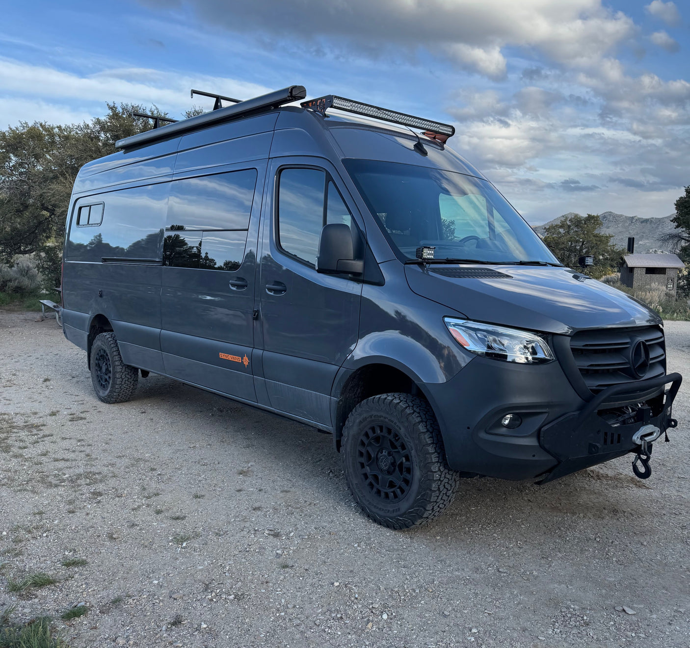 Dark gray Mercedes Sprinter rv rental camper van with off-road tires and light bars, parked on a gravel path under a cloudy sky.