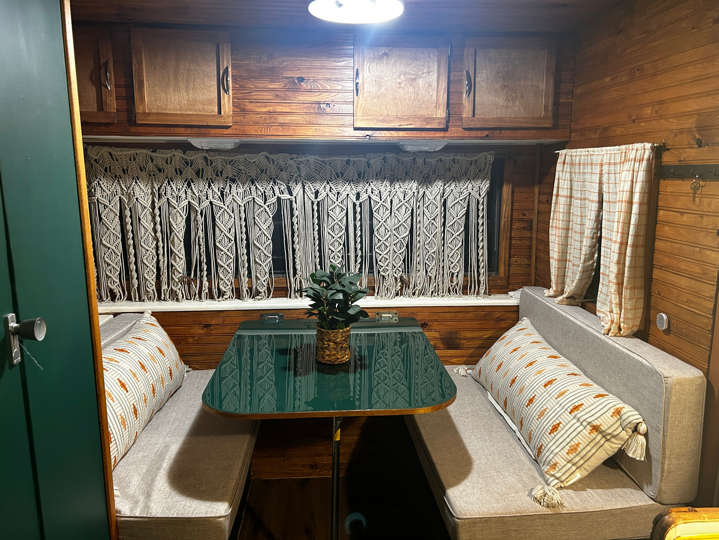 Inside a charming RV rental: Wood-paneled walls, a green dinette table, cozy seating, and macrame window decor.