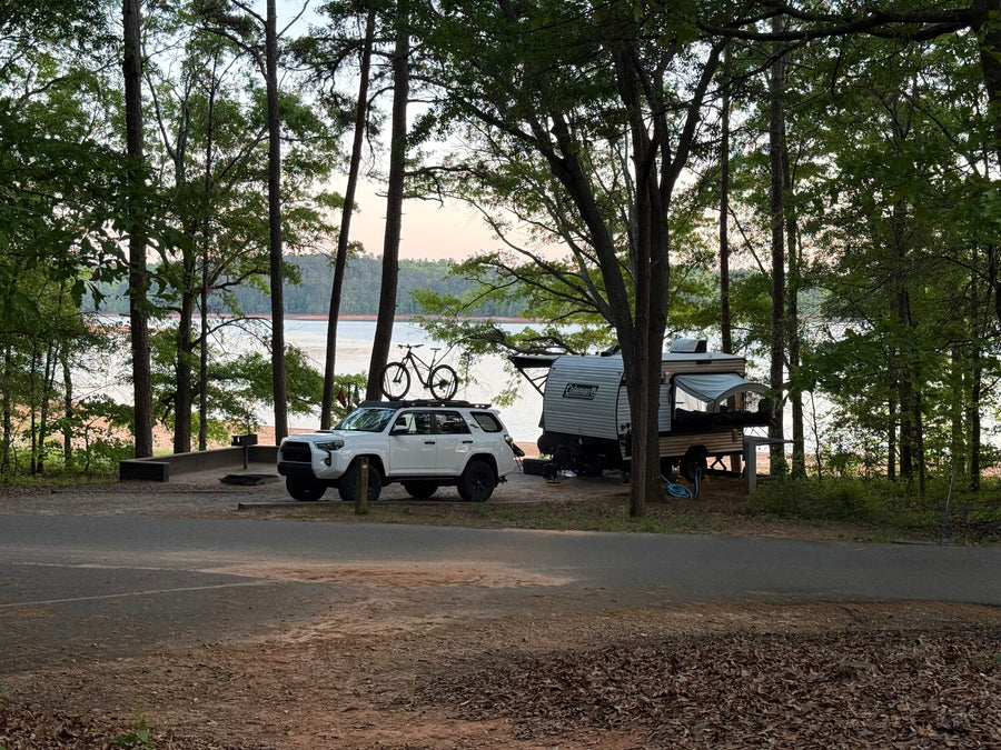 An RV rental and white SUV with a bike on top parked at a scenic lakeside campsite among tall trees.|This Coleman travel trailer