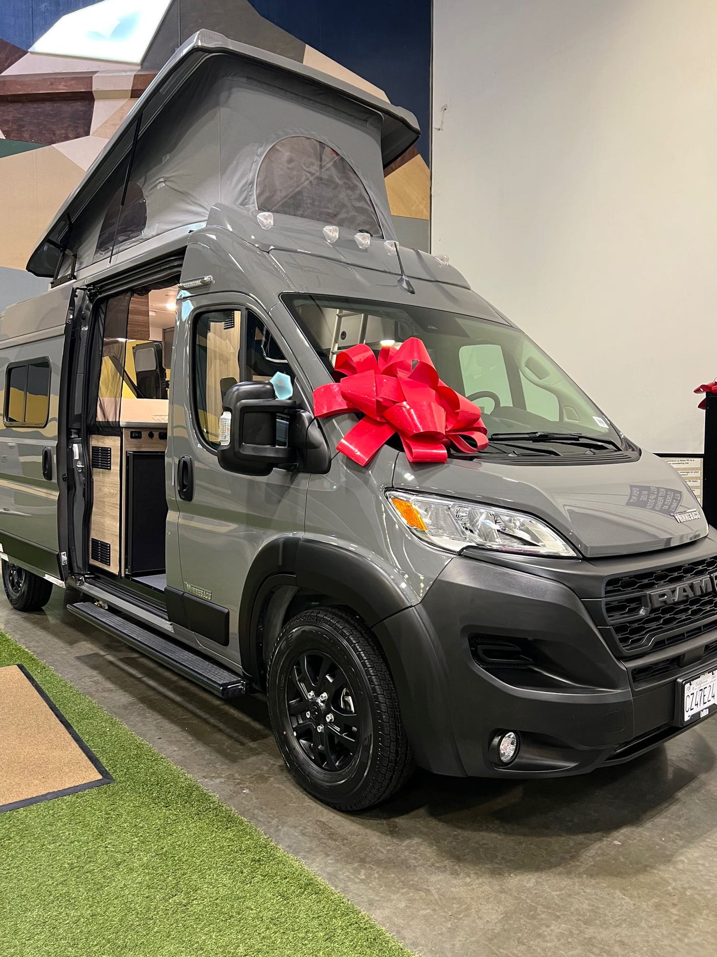 Grey Winnebago Ram camper van, featuring a pop-top and red bow, showcased for potential RV rental. Interior is visible.