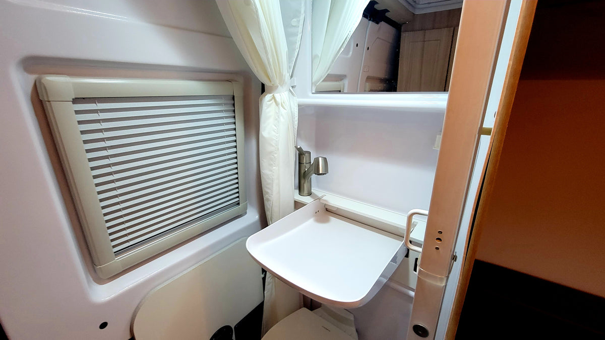 View inside a compact RV rental bathroom, featuring a sink, pull-out counter, toilet, and window with blinds. — photo 15