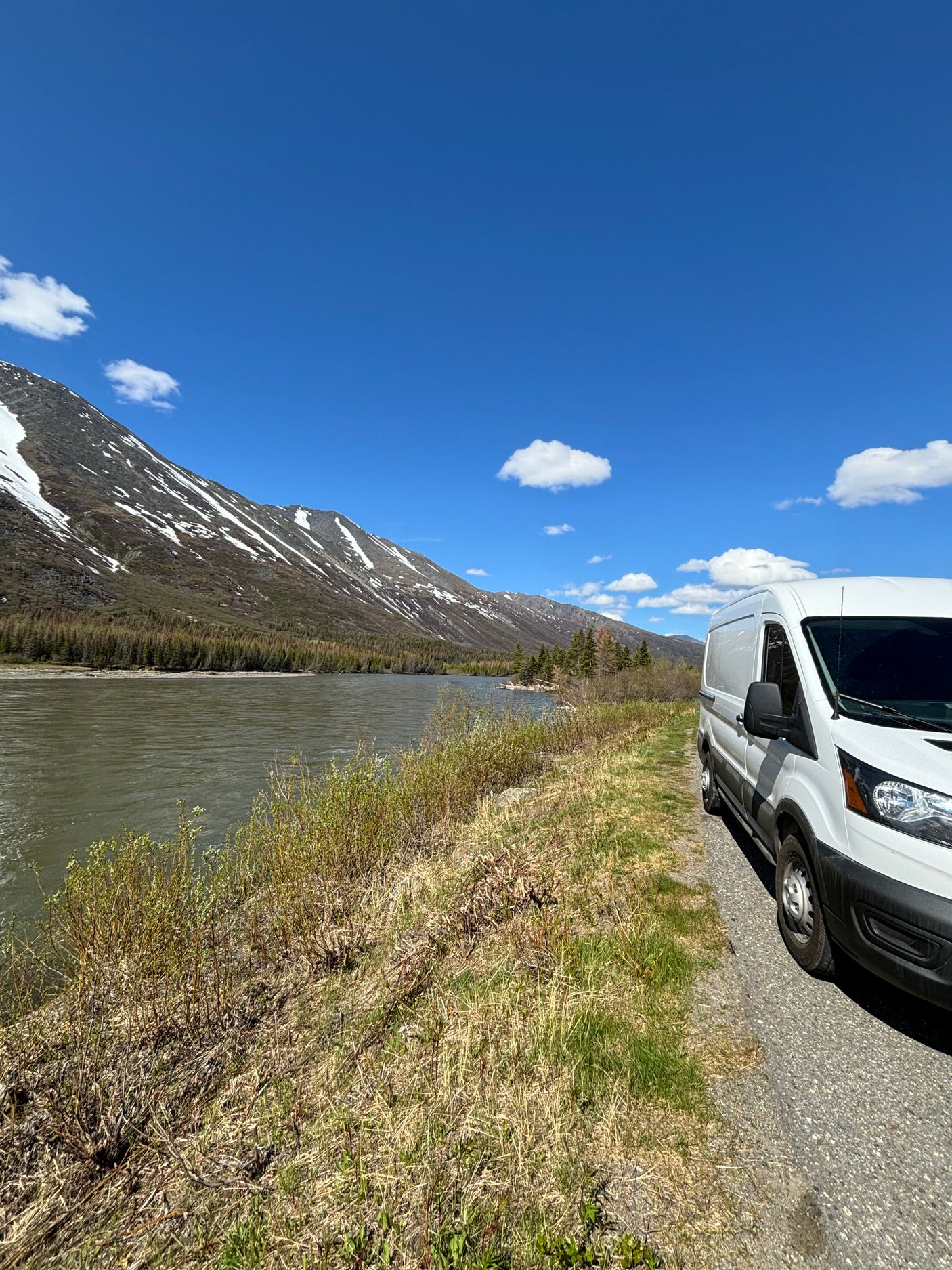 RV rental parked beside a scenic river with snowy mountains under a clear blue sky.