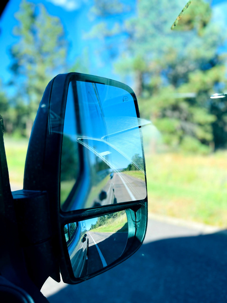 View from an RV rental: Open road ahead reflected in the passenger side mirror, showing highway and distant trees.