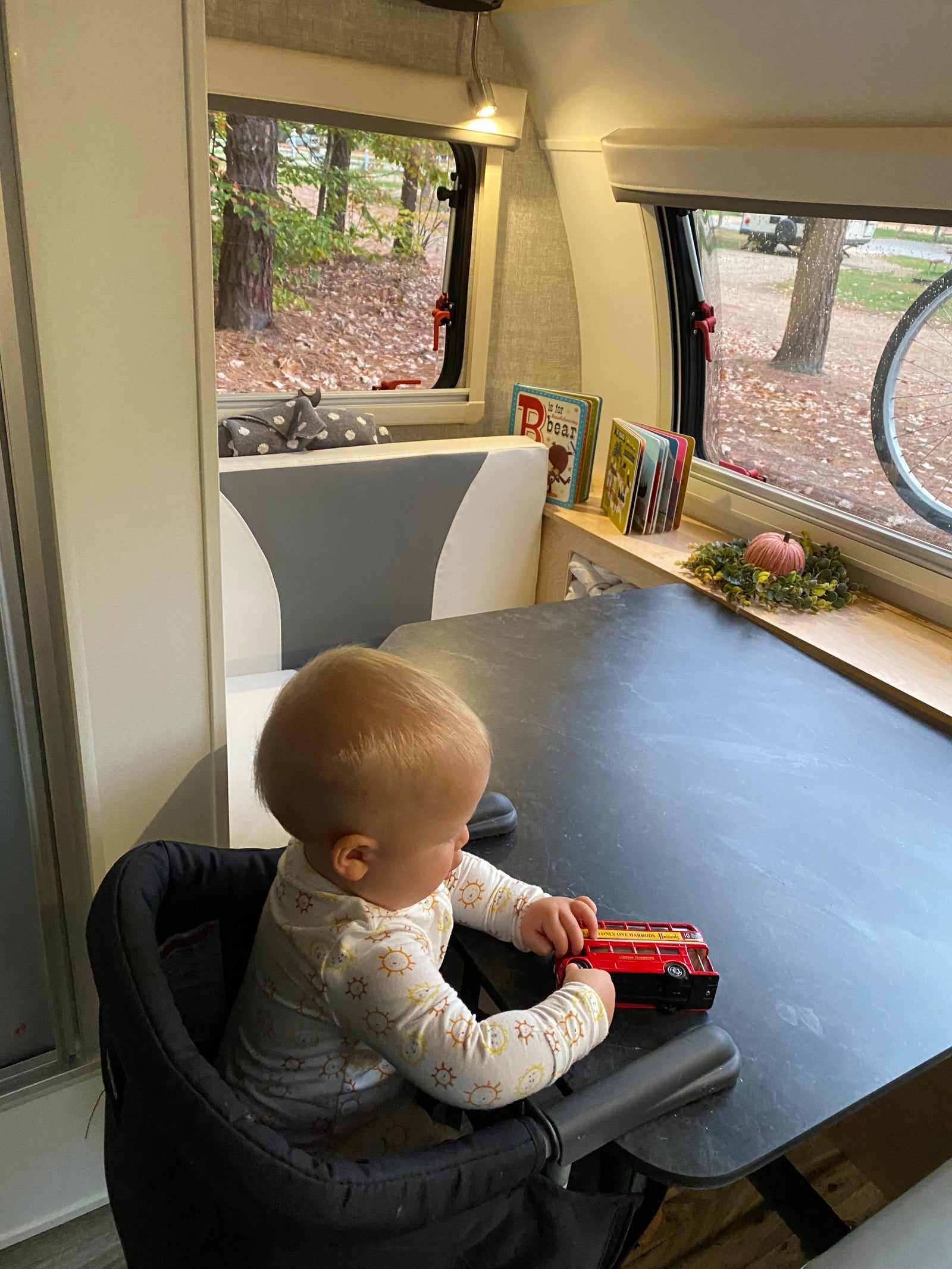 A baby plays with a red toy bus at a table inside an RV rental, with autumn foliage visible outside the window.