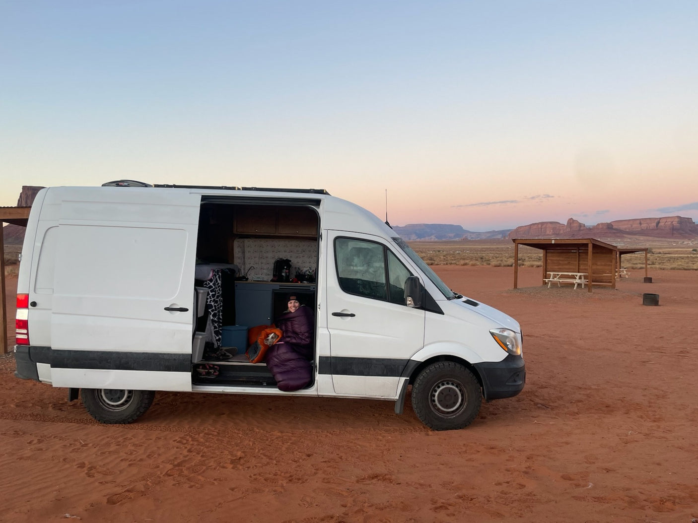 White RV rental parked in desert. Person and dog cozy in sleeping bag, open van door. Mesas under dawn/dusk sky.