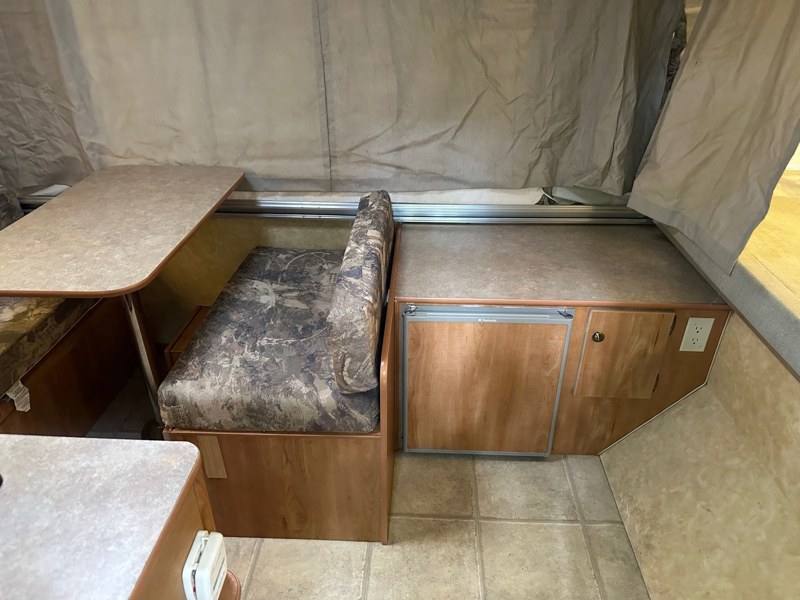 is set up on a gravel campsite amidst green trees.|Open door of an RV rental pop-up camper. See its interior with tiled floor & exterior canvas tent at a campsite.|Inside an RV rental: a cozy pop-up camper dinette with a camo bench