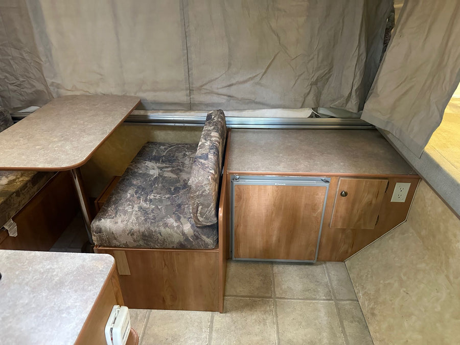 is set up on a gravel campsite amidst green trees.|Open door of an RV rental pop-up camper. See its interior with tiled floor & exterior canvas tent at a campsite.|Inside an RV rental: a cozy pop-up camper dinette with a camo bench