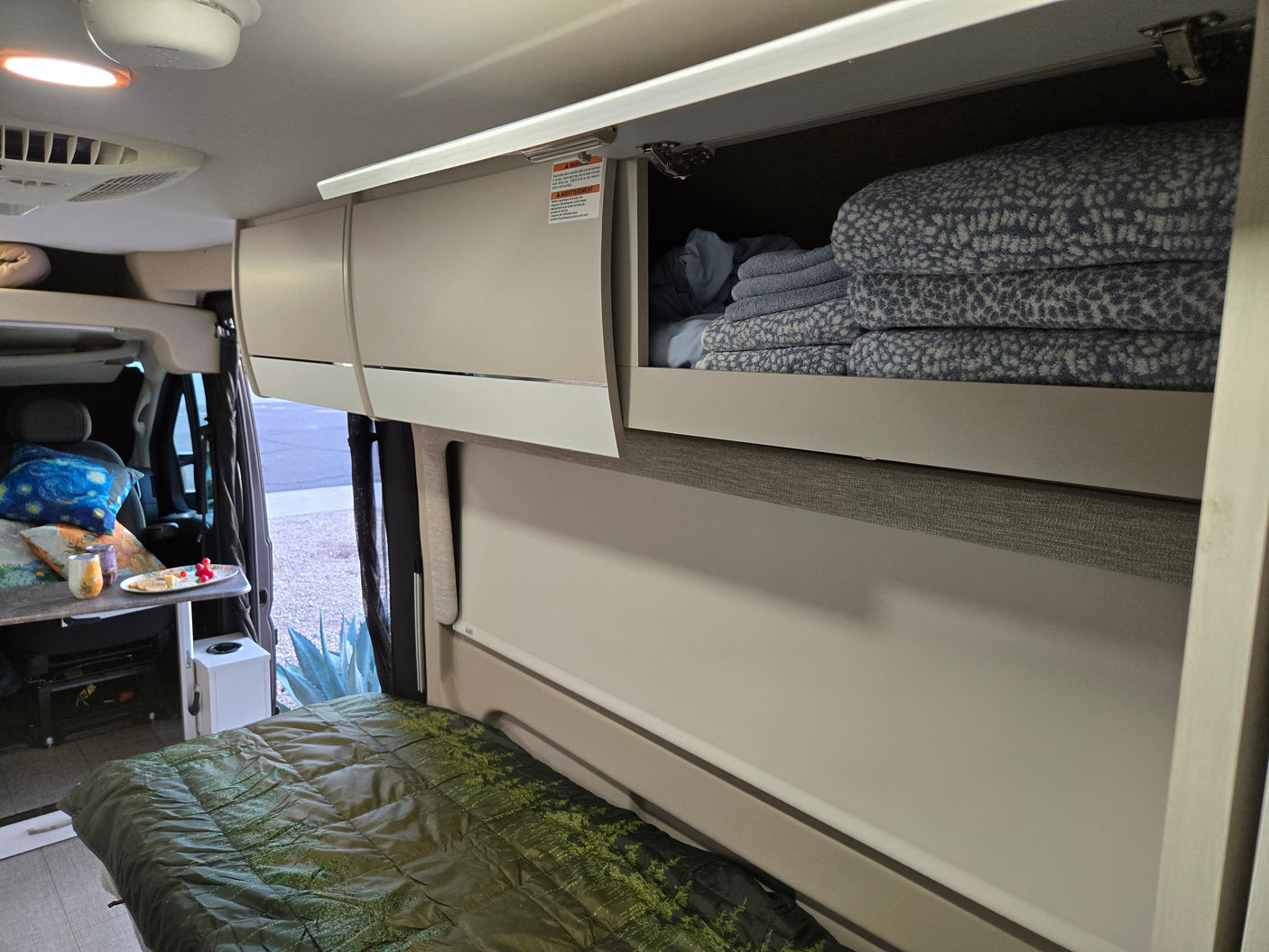 Cozy rv rental interior: open cabinet with blue towels, a green bed, and snacks on a side table. Ready for comfortable travel.