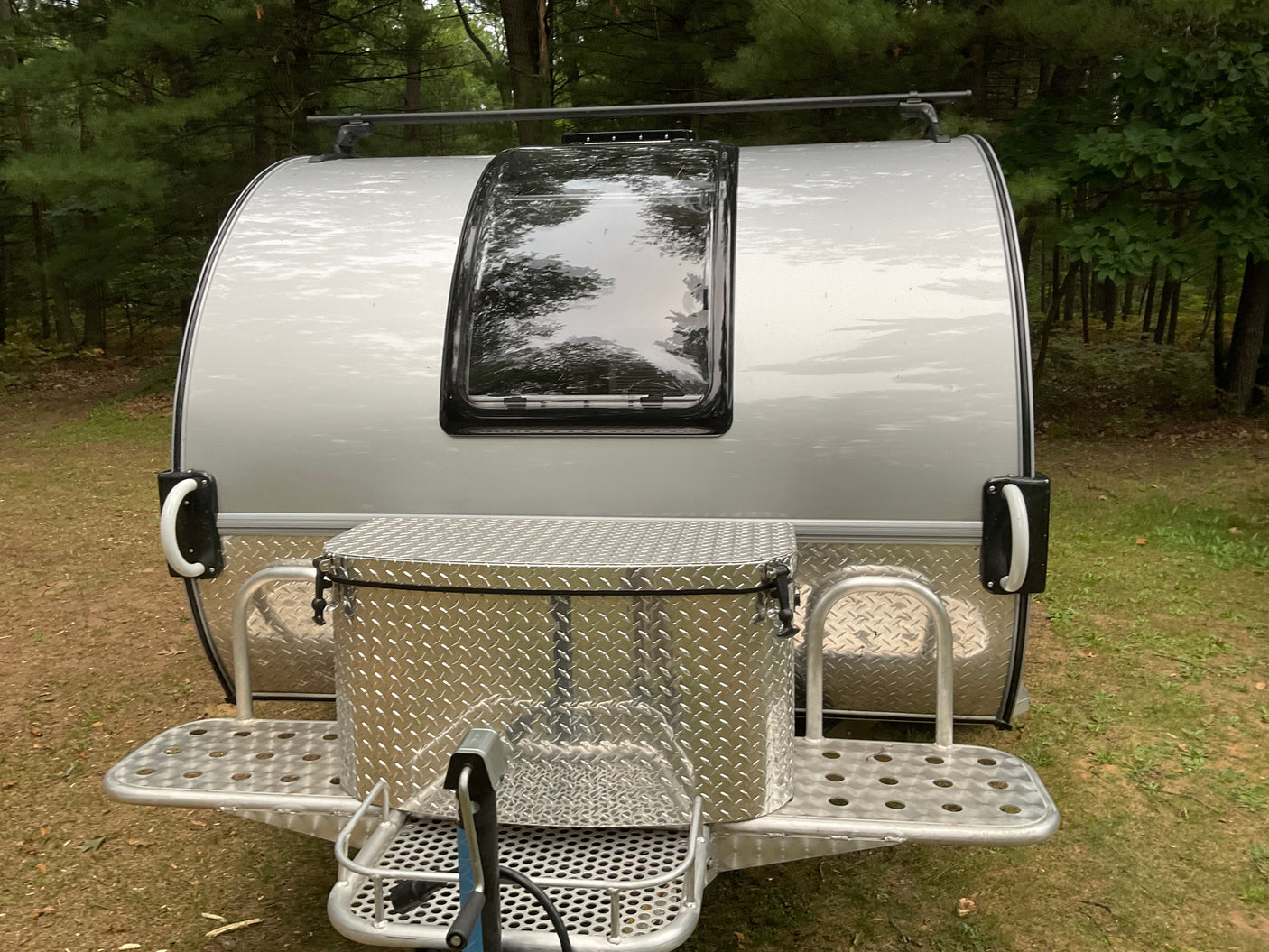 Silver teardrop travel trailer (RV rental) with a large front storage box and roof rack, parked in a wooded area.