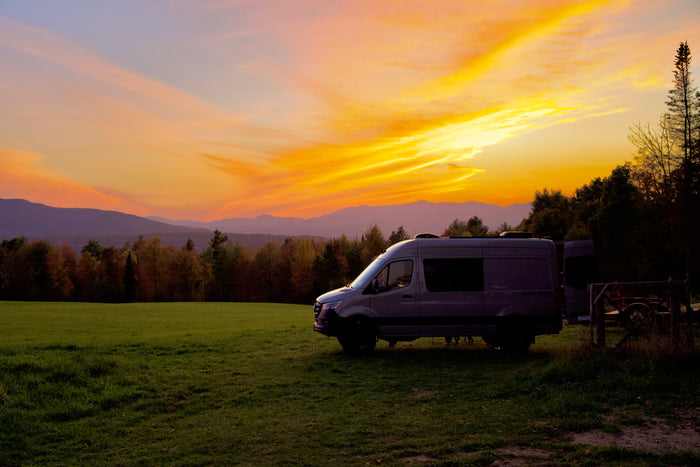 An RV rental camper van bathed in the golden light of a stunning sunset over mountains and a field. Travel dreams come true!