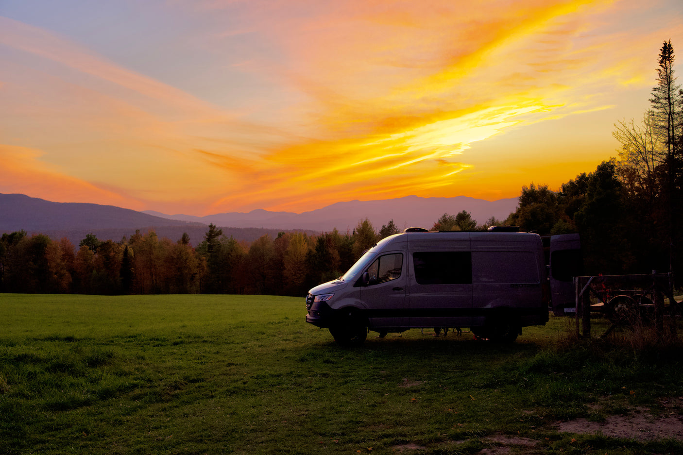 An RV rental camper van bathed in the golden light of a stunning sunset over mountains and a field. Travel dreams come true!