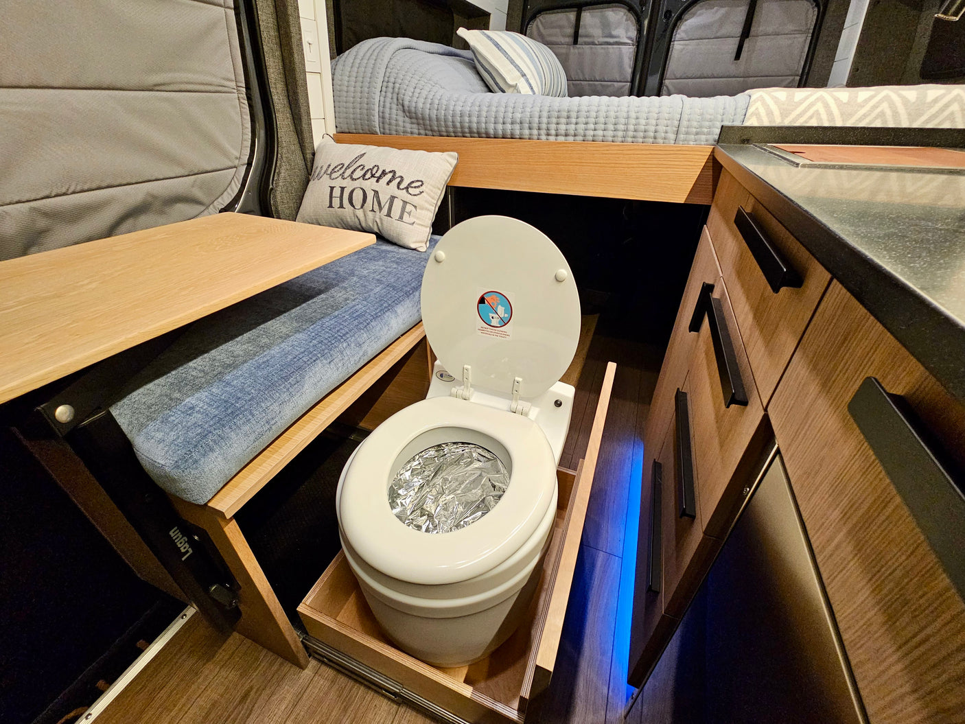 This compact rv rental interior shows a pull-out toilet, a bed, a wooden table, and storage cabinets.