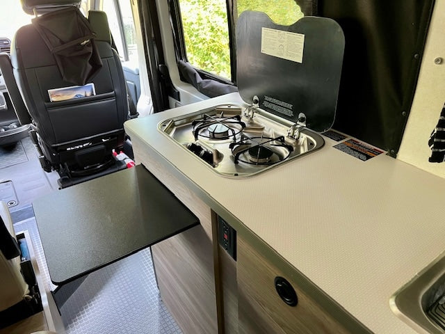 RV rental interior: compact kitchen with two-burner stove, sink, and pull-out table. Front seat storage visible for road trips.