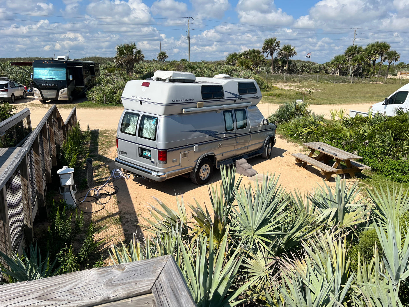 RV rental: Airstream 190 camper van at a sandy Florida campsite by the beach, surrounded by palm trees and a picnic table.