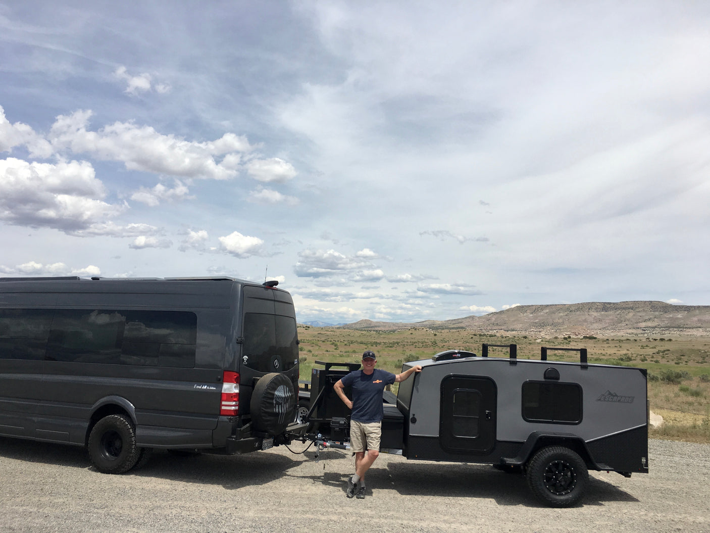 Man smiles by a dark grey RV rental van and compact trailer, ready for adventure in a vast, cloudy landscape.