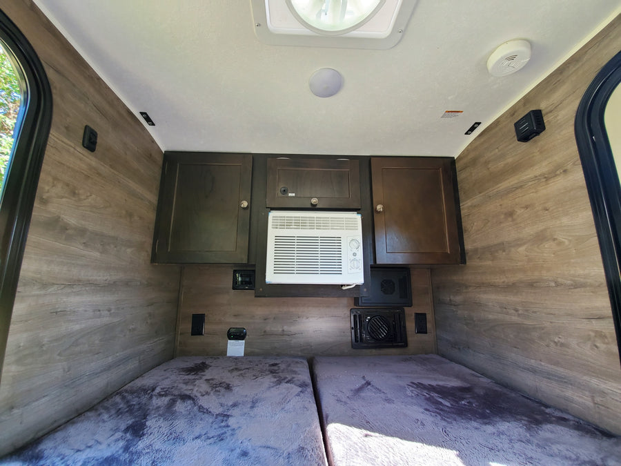 RV rental interior: wood-paneled walls, twin beds, dark cabinets, AC unit, & ceiling fan. Cozy camper space.