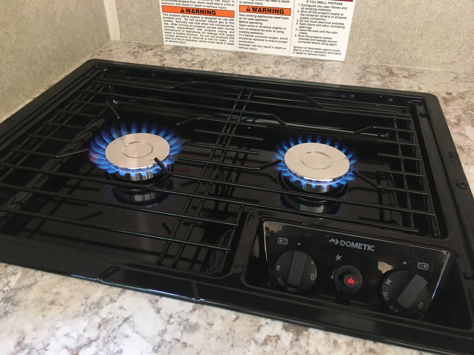 An RV rental gas stovetop with two blue-flamed burners lit, featuring a Dometic control panel and important warning labels.