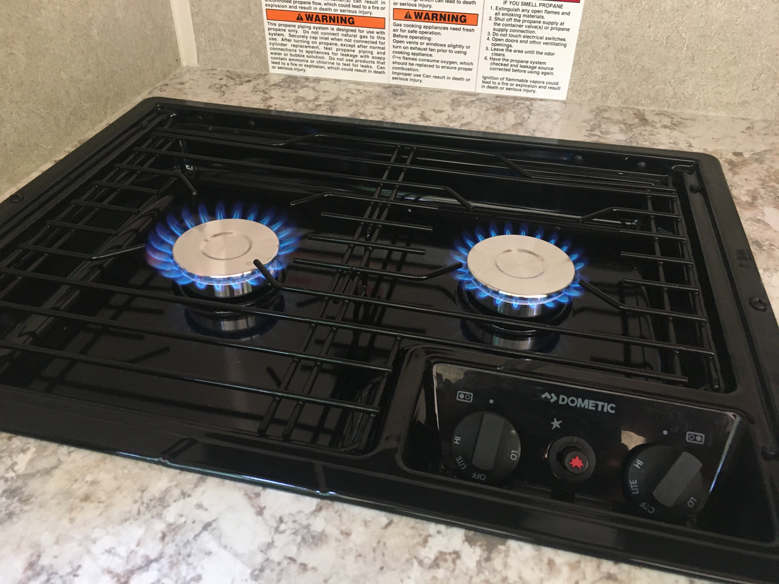 An RV rental gas stovetop with two blue-flamed burners lit, featuring a Dometic control panel and important warning labels.