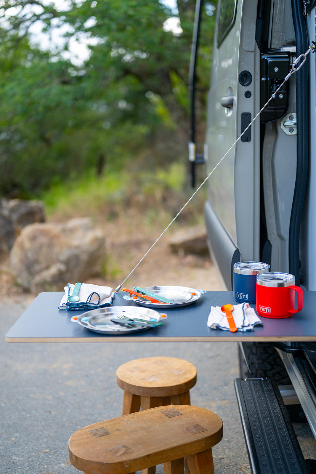 RV rental van's outdoor dining setup: a portable table with plates, colorful cutlery, YETI mugs, and wooden stools.