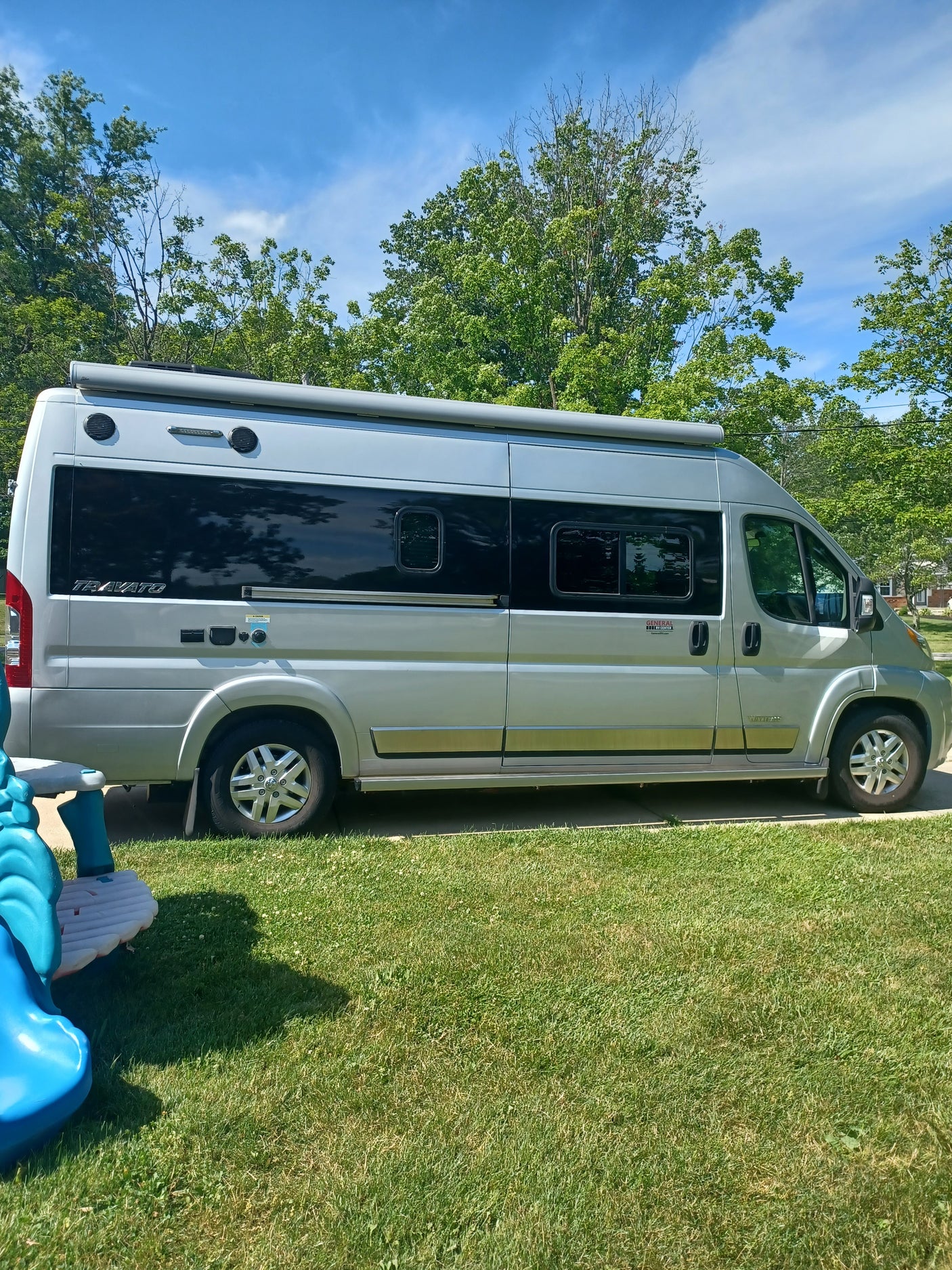 A Winnebago Travato camper van, ideal as an rv rental, parked on a sunny driveway beside green grass and trees.