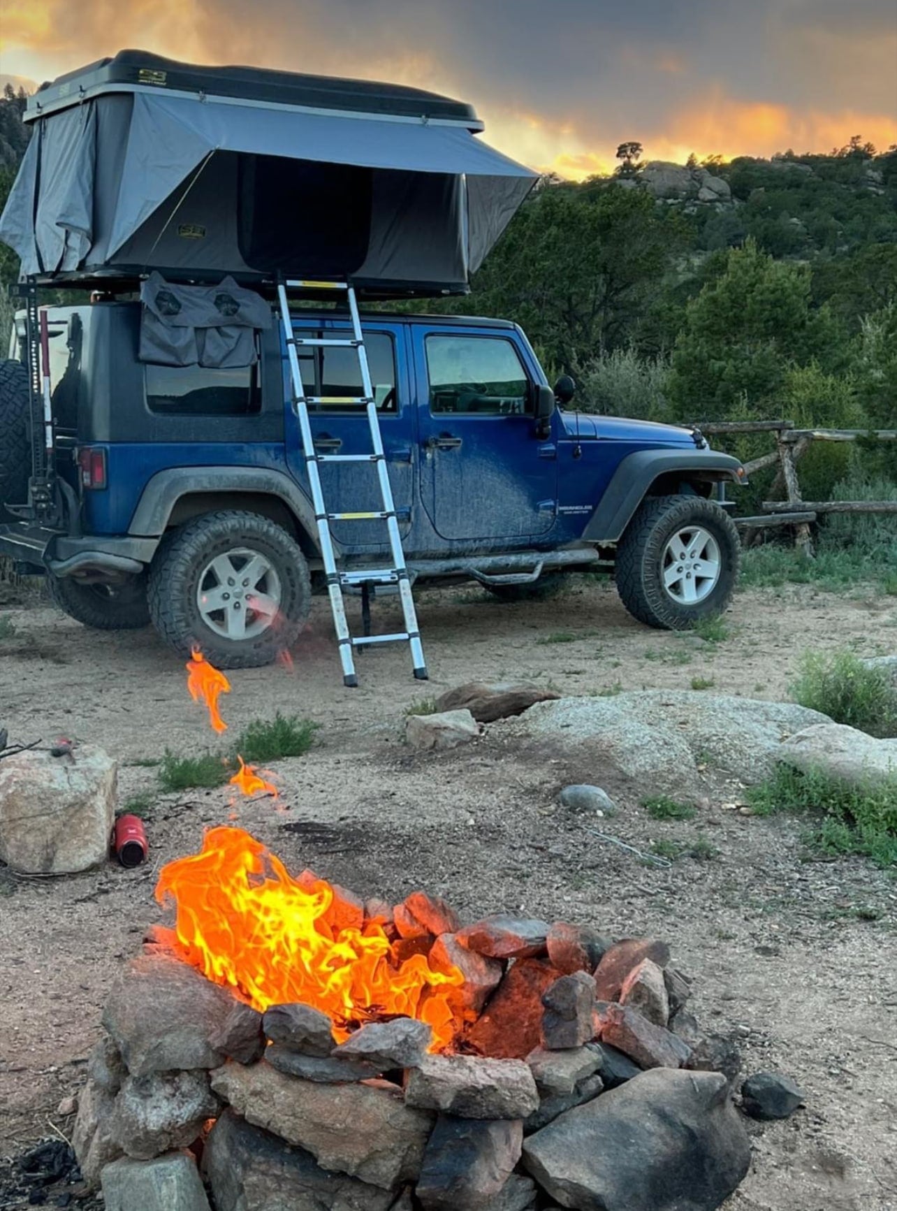like an RV rental for rugged exploration.|This blue Jeep with a deployed rooftop tent by a lake offers an amazing RV rental experience amidst scenic mountains.|A blue Jeep Wrangler with an open rooftop tent parked in a sun-dappled forest campsite — photo 2