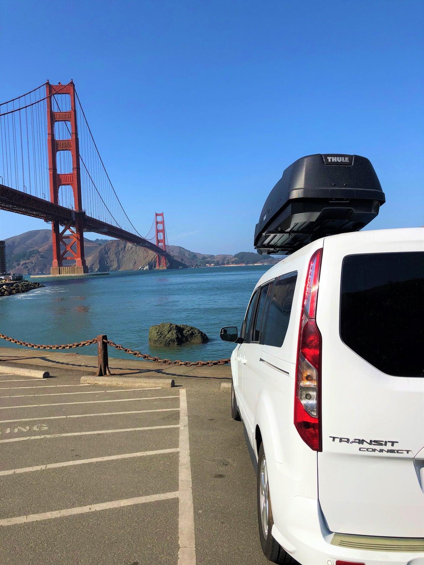 White van, possibly an RV rental, with a Thule roof box, parked near the Golden Gate Bridge and bay under a clear sky.