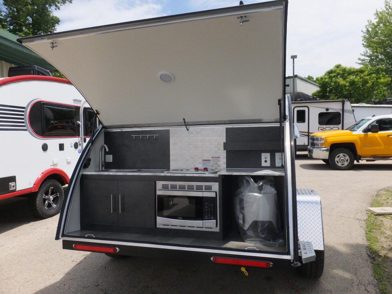 parked on a driveway at dusk.|Compact dark grey teardrop RV rental and other campers on display at a dealership.|Compact silver and black teardrop RV for rental
