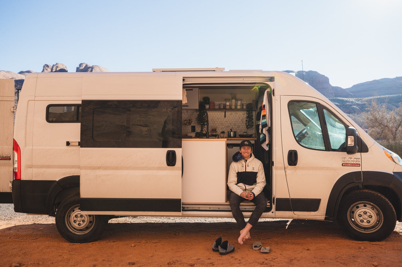 A man smiles from his custom white camper van's open door, showcasing its interior against a desert backdrop. Ideal RV rental.