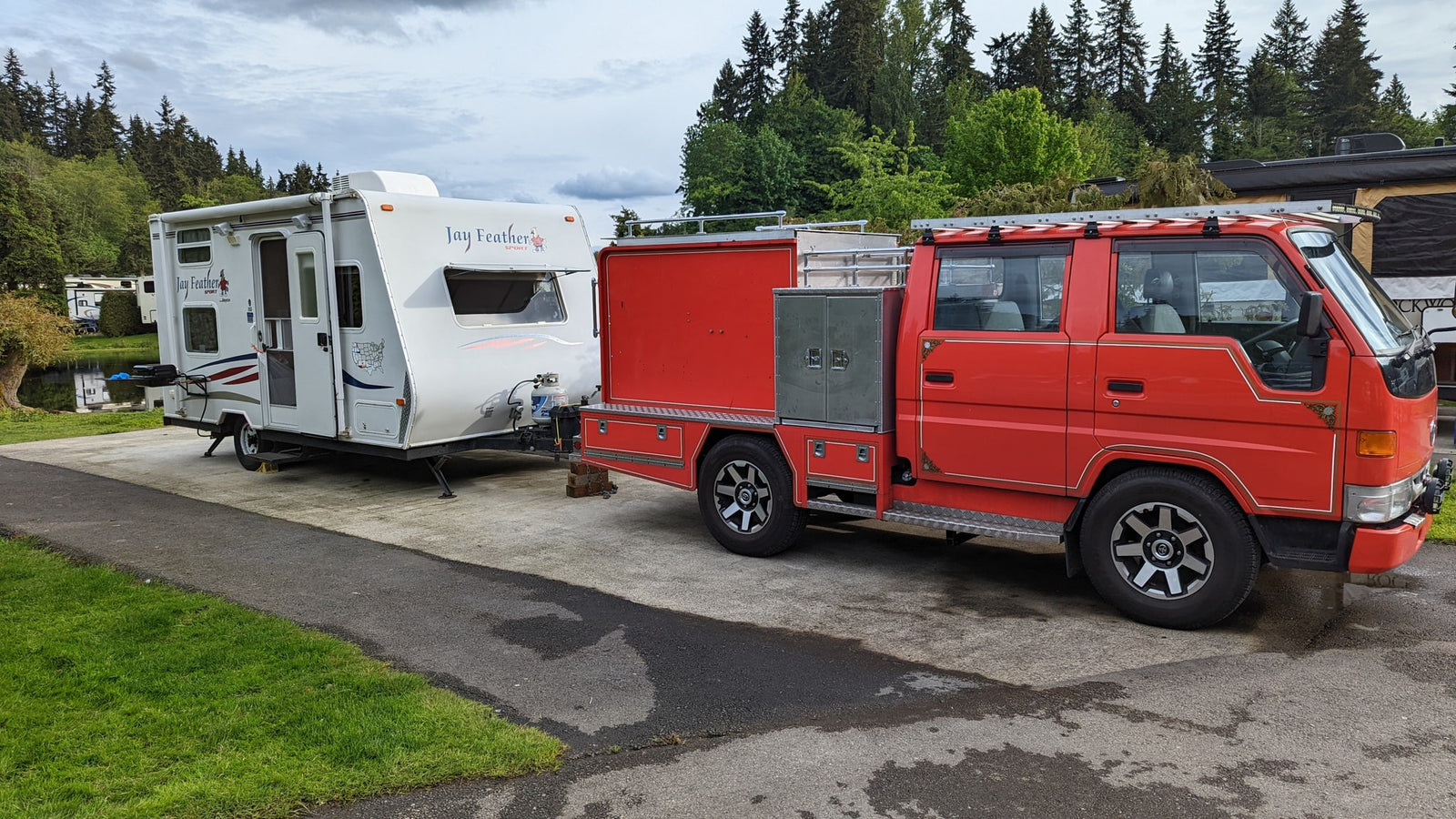 Red custom truck towing a white Jay Feather travel trailer in a green, scenic campground, ideal for an RV rental.