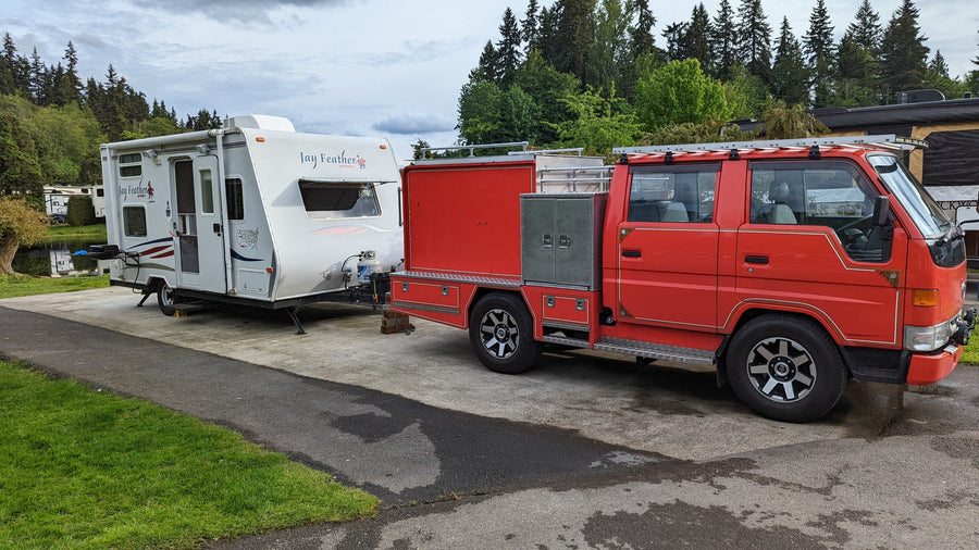 Red custom truck towing a white Jay Feather travel trailer in a green, scenic campground, ideal for an RV rental.