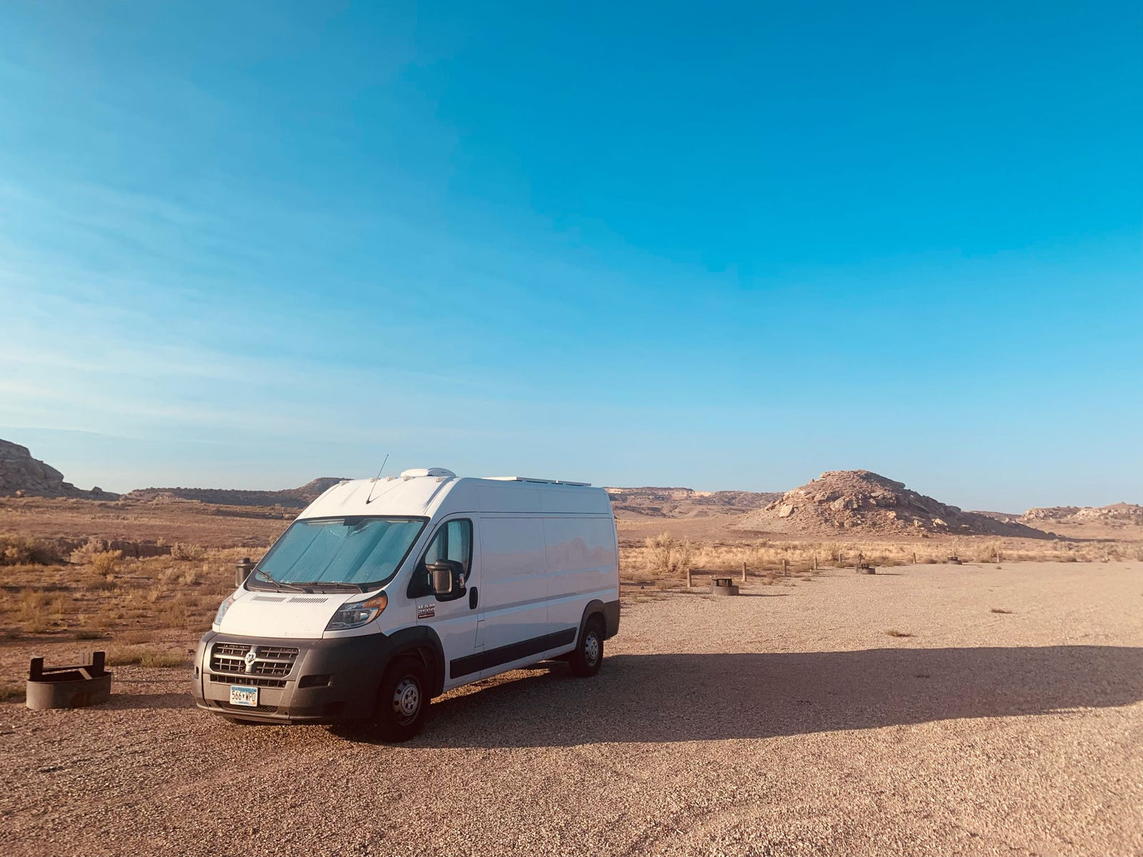A white Ram ProMaster RV rental campervan is parked in a desert campsite with rocky hills under a blue sky.|Cozy RV rental interior: wood-paneled ceiling