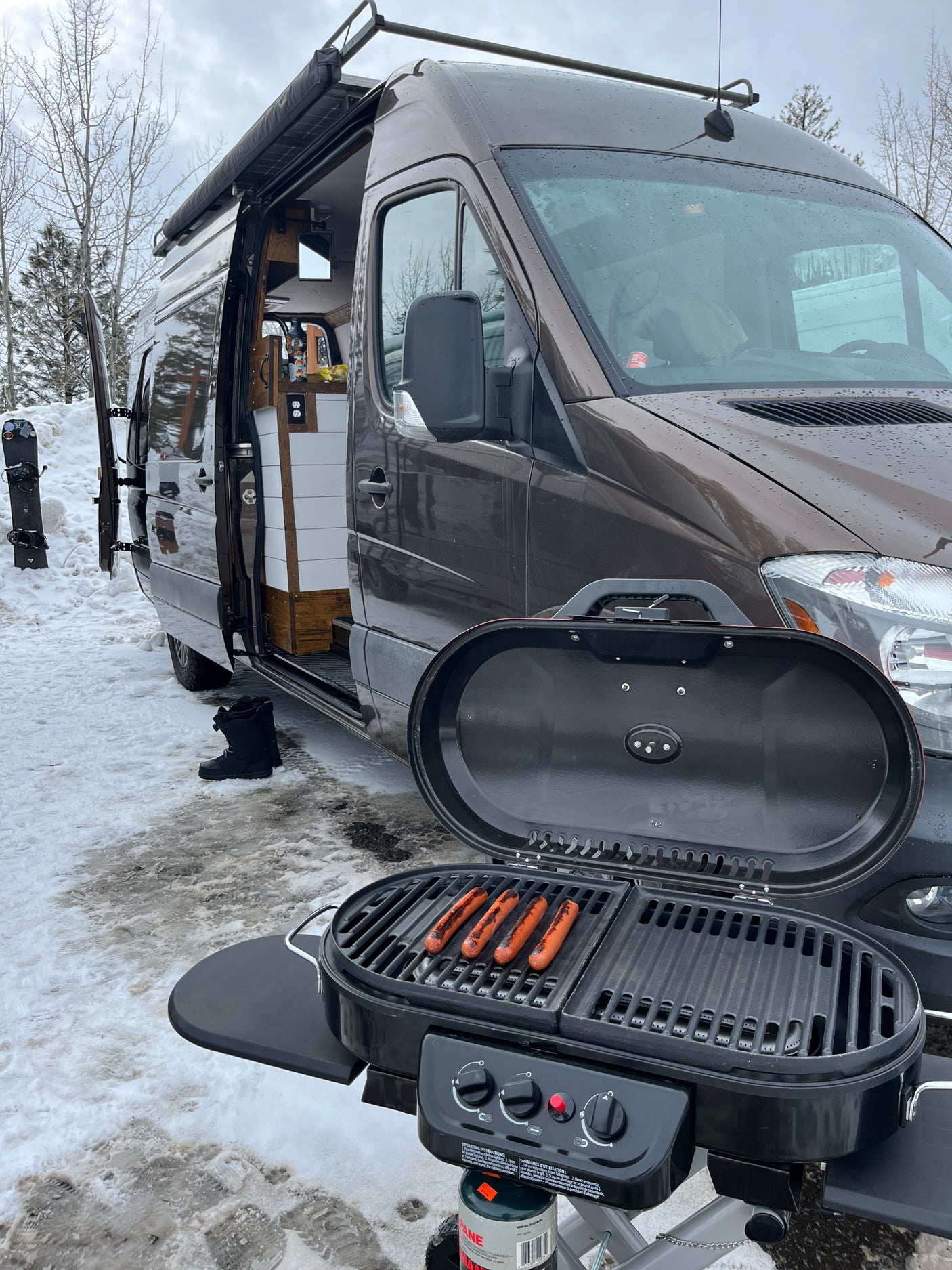 An RV rental van parked in snow, with a grill cooking hotdogs outside. Perfect for winter camping trips.