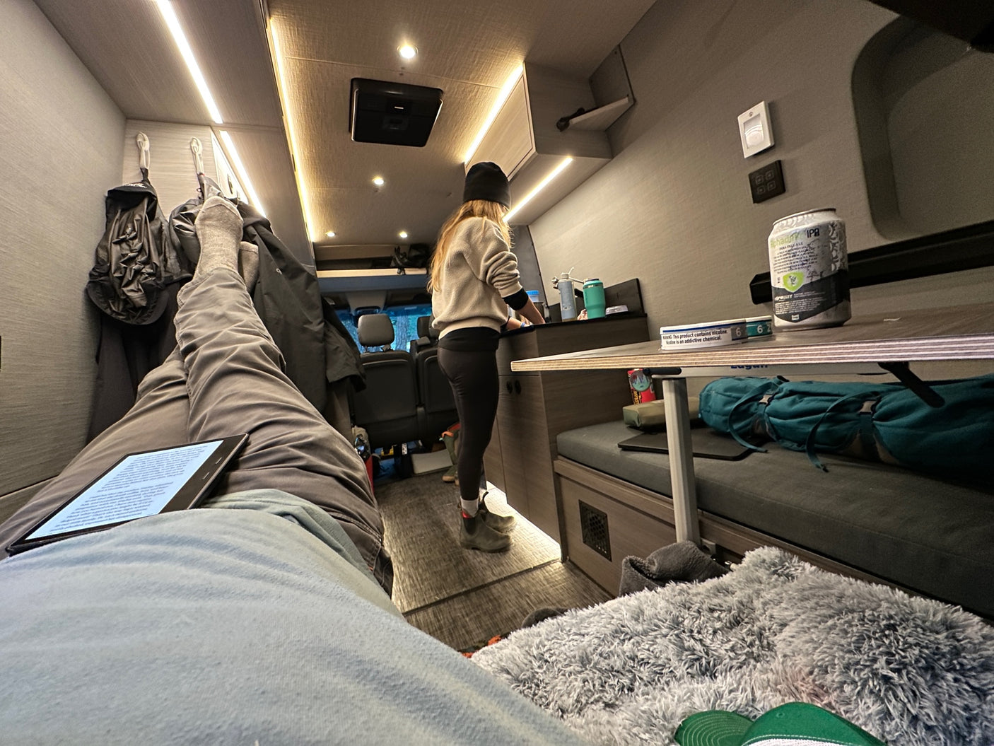 Cozy RV rental interior: one person cooks, while another relaxes on a bench with a Kindle and legs up, enjoying van life.