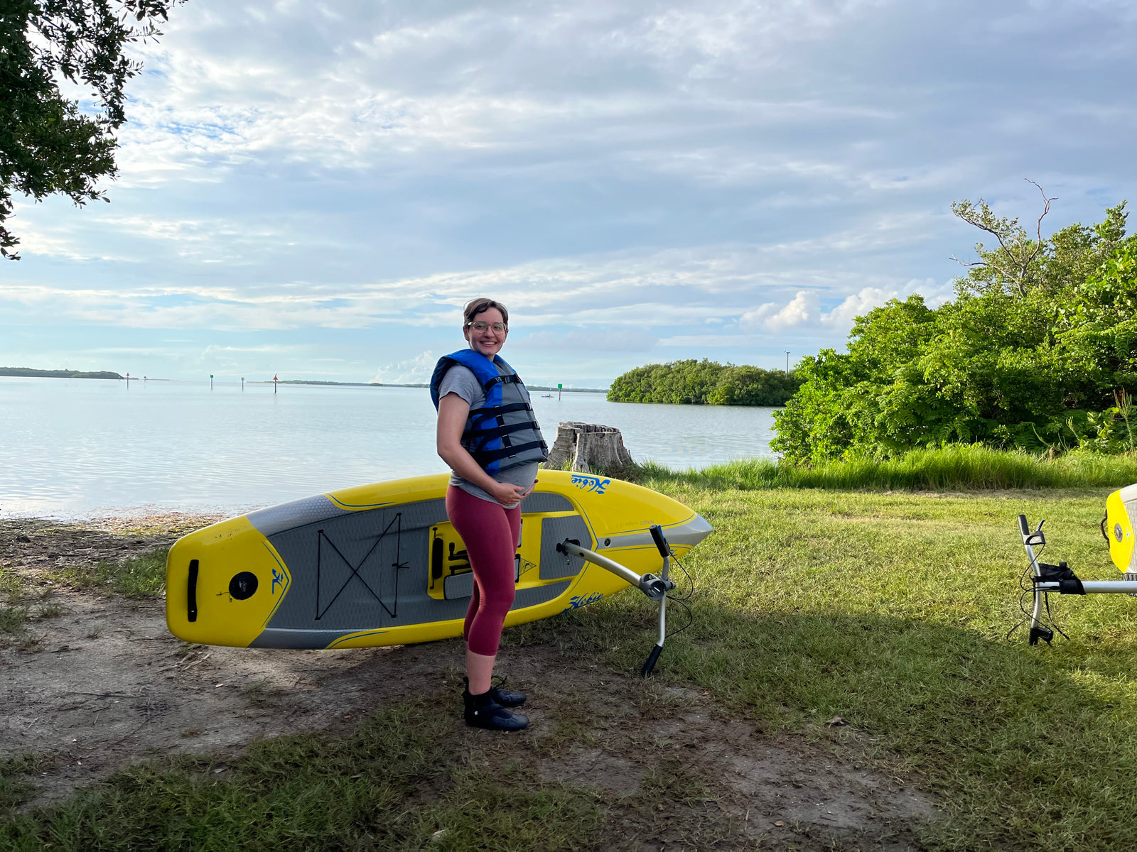 Smiling pregnant woman in a life vest by a yellow Hobie SUP near calm water and green banks. Enjoy nature with an RV rental!