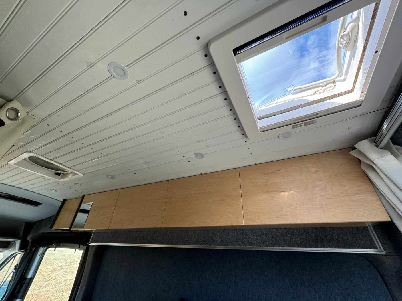RV rental interior: ceiling with recessed lights, large skylight, and wood overhead cabinets in a modern camper van design.