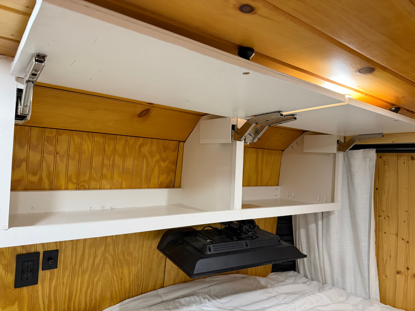 RV rental interior shows opened overhead cabinets, wood paneling, a mounted TV, and electrical outlets near the bed.