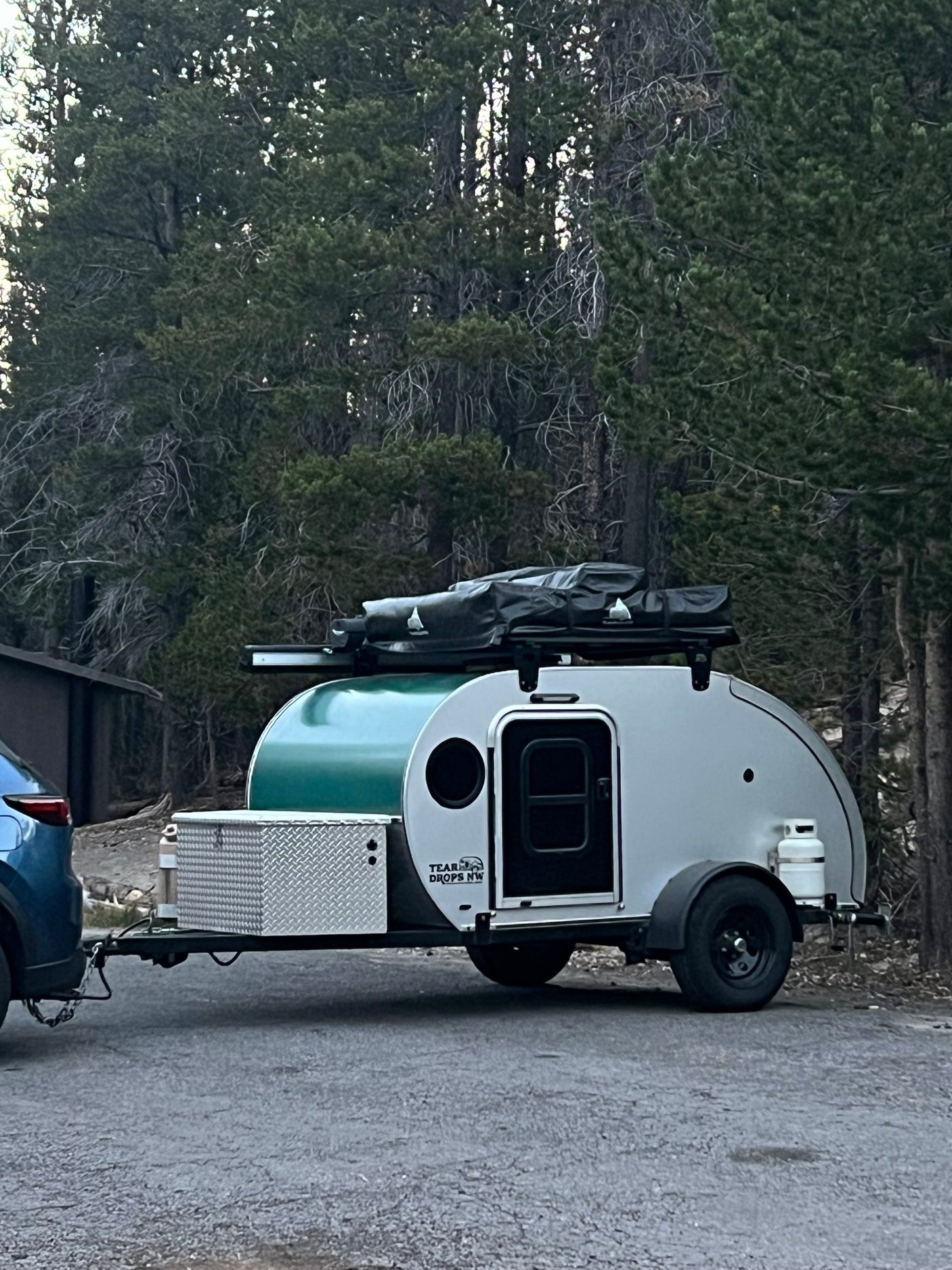 Compact teardrop RV rental attached to a blue SUV, parked on pavement in a forest setting. Equipped for outdoor camping.