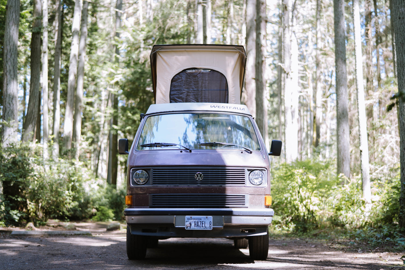 Front view of a Westfalia camper van, perfect for rv rental, parked in a sunny forest with its pop-top extended.