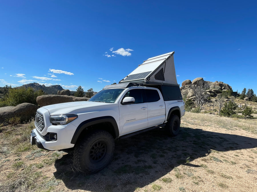 White Toyota Tacoma RV rental with pop-top camper & solar panels, parked in a rugged mountain setting.