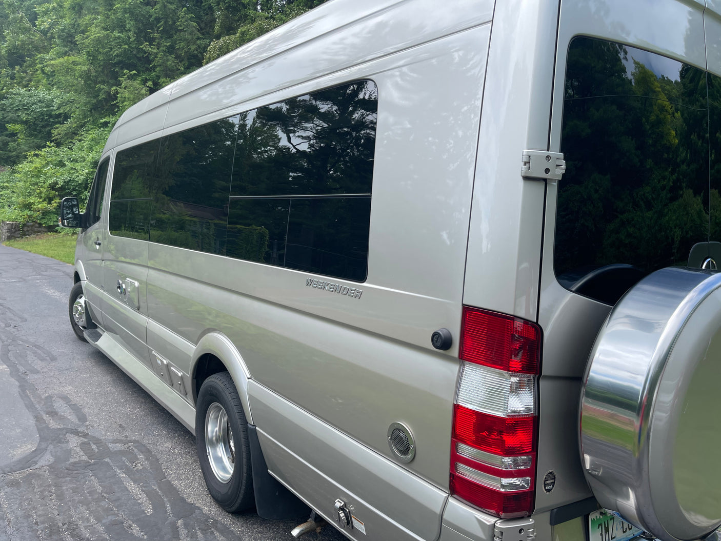 Silver Mercedes-Benz Sprinter Weekender RV for rental, showing its spacious side and rear with a spare tire carrier, parked by trees.