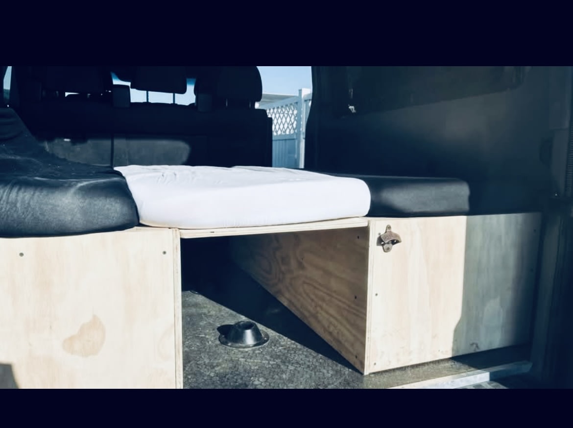 Inside an RV rental van: a cozy bed, wooden storage with a bottle opener, and a dog bowl below. Ready for adventures!