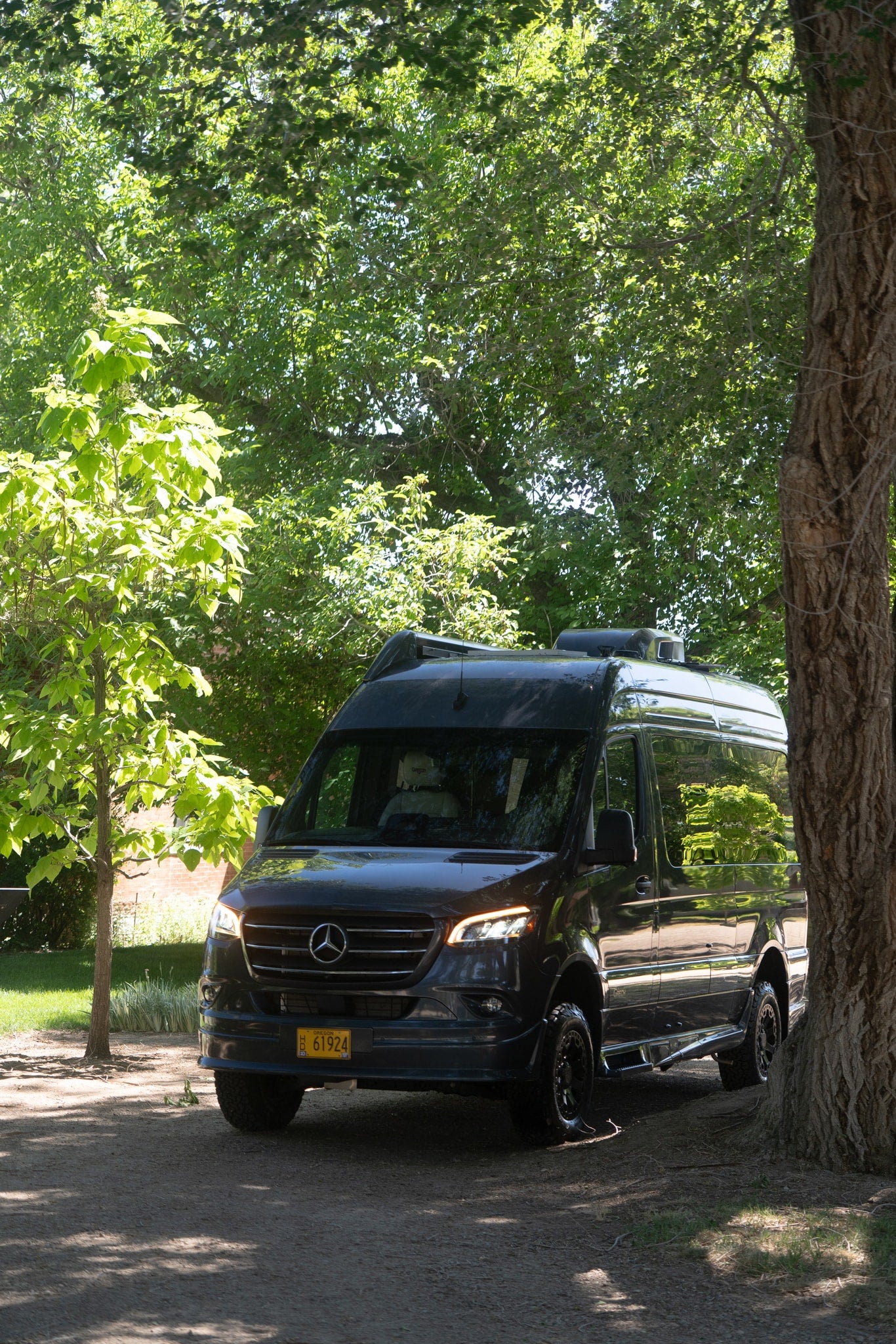 Dark gray Mercedes-Benz Sprinter rv rental van parked under dappled sunlight from lush green trees, ready for adventure. — photo 19