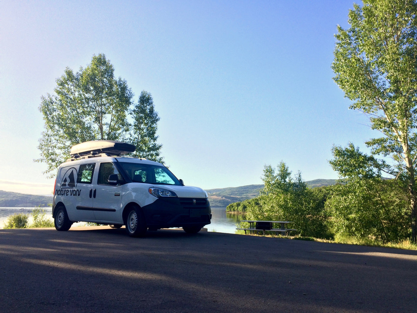 White "Nature Vans" RV rental parked by a tranquil lake, green trees, and mountains under a clear blue sky.