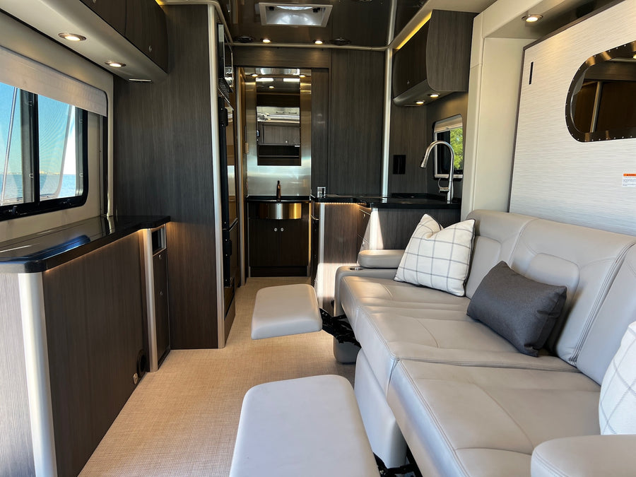 Step into this sleek rv rental featuring a comfy grey leather sofa, footrests, modern kitchen, and a window with a water view.